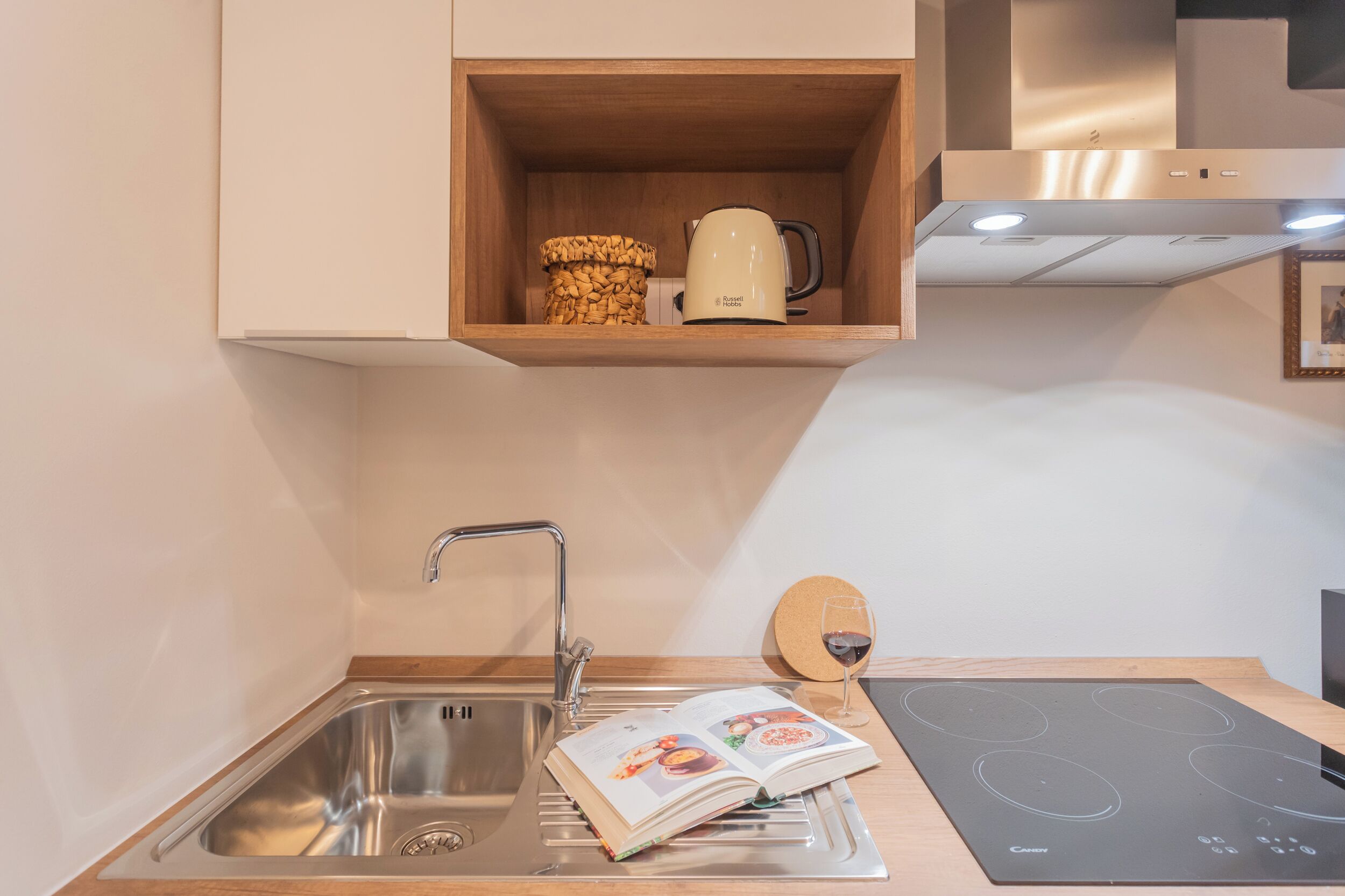 Una cocina moderna con un lavamanos de acero inoxidable, una placa de inducción integrada y un estante de madera sobre el lavamanos con una cafetera y una cesta de mimbre.