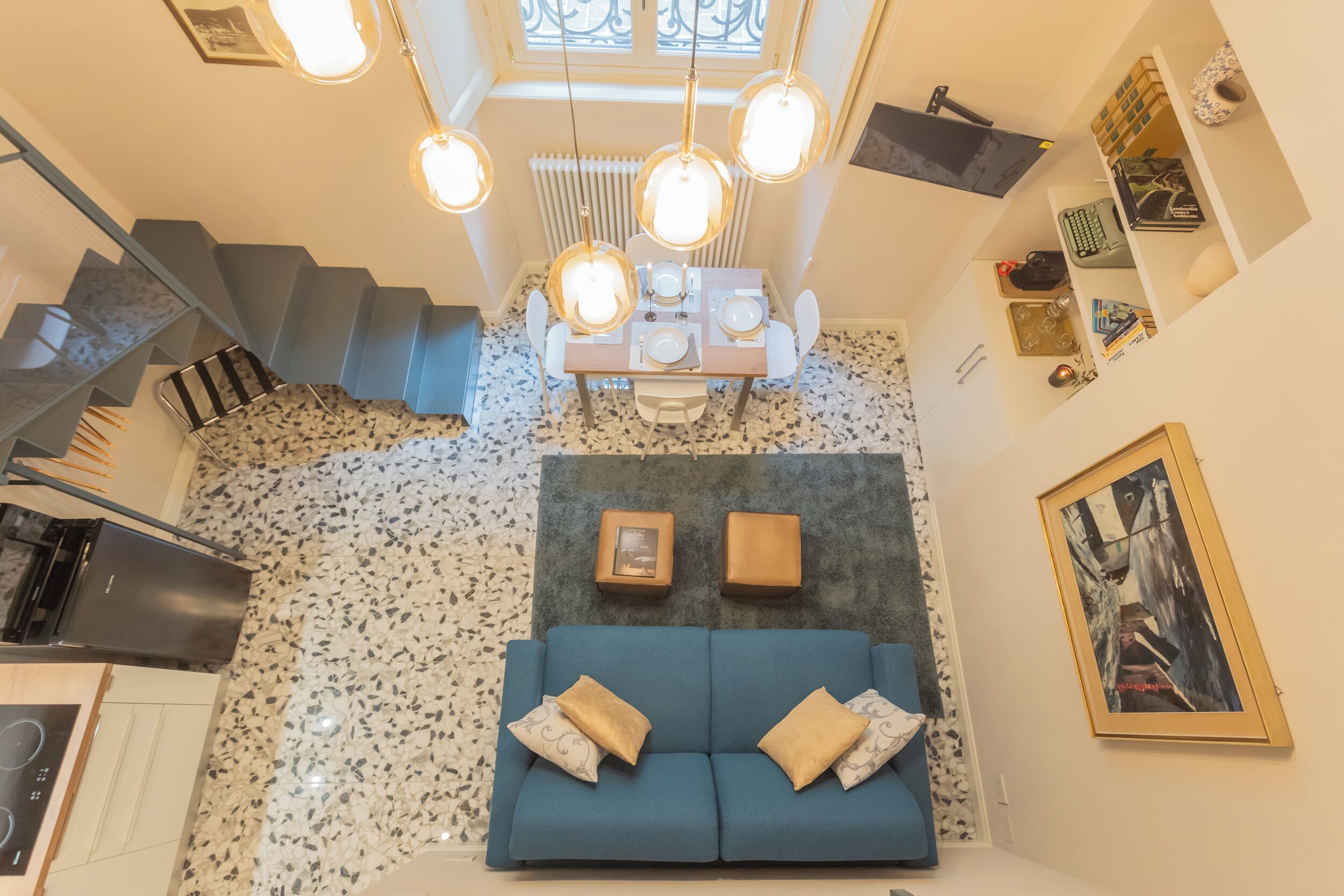 A modern kitchen with a built-in stove, a dining area with a table and chairs, a blue sofa with cushions, and a staircase leading to an upper floor.