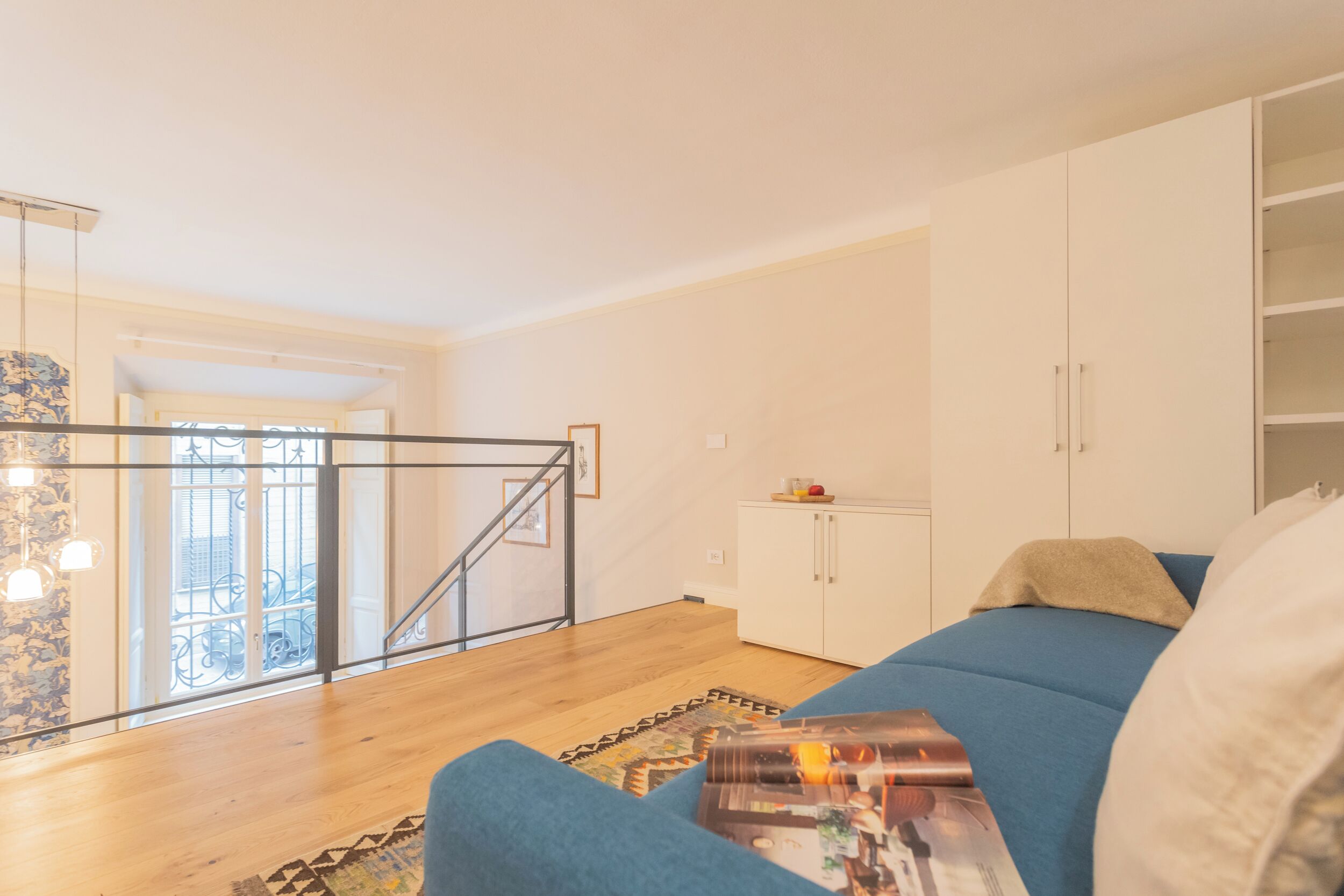 A modern living room with a blue sofa, a coffee table with magazines, a white cabinet, and a staircase leading to an outdoor balcony.