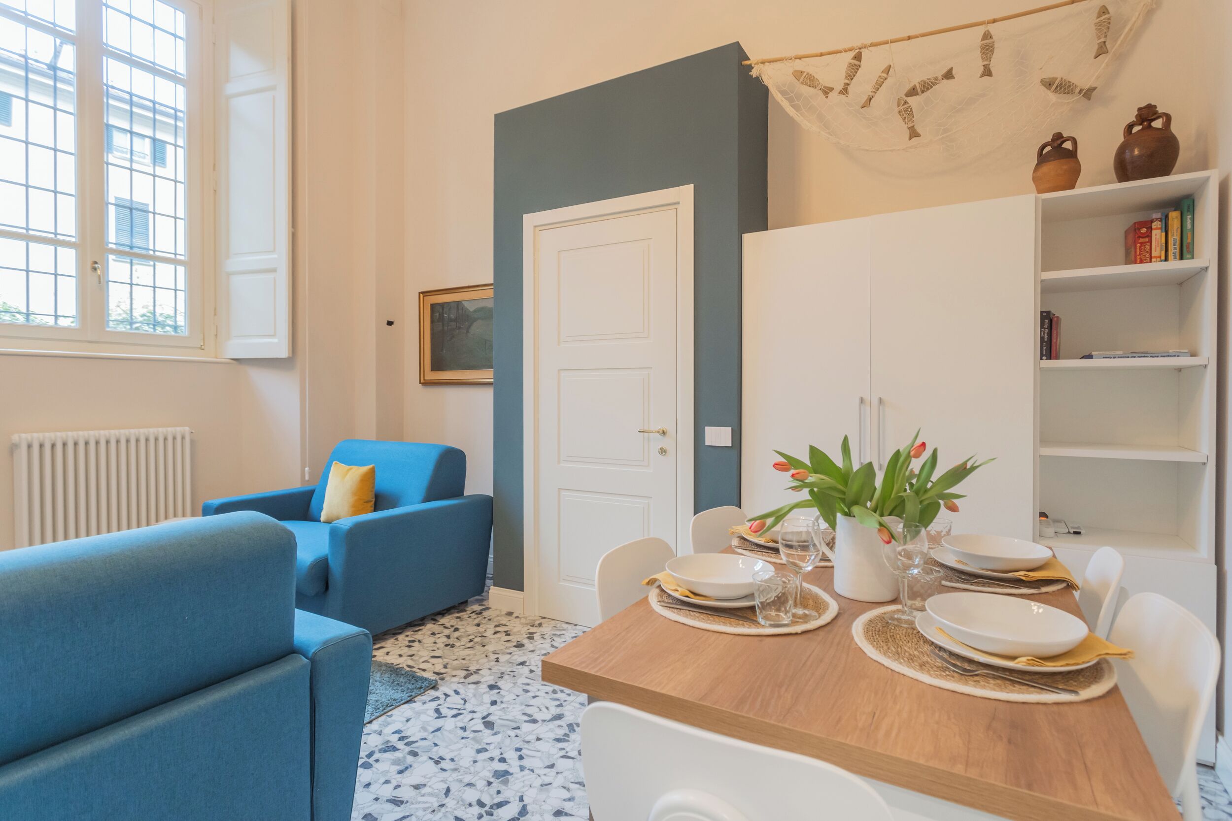 Un coin repas avec une table en bois dressée pour quatre personnes, des chaises blanches et un canapé bleu en arrière-plan.