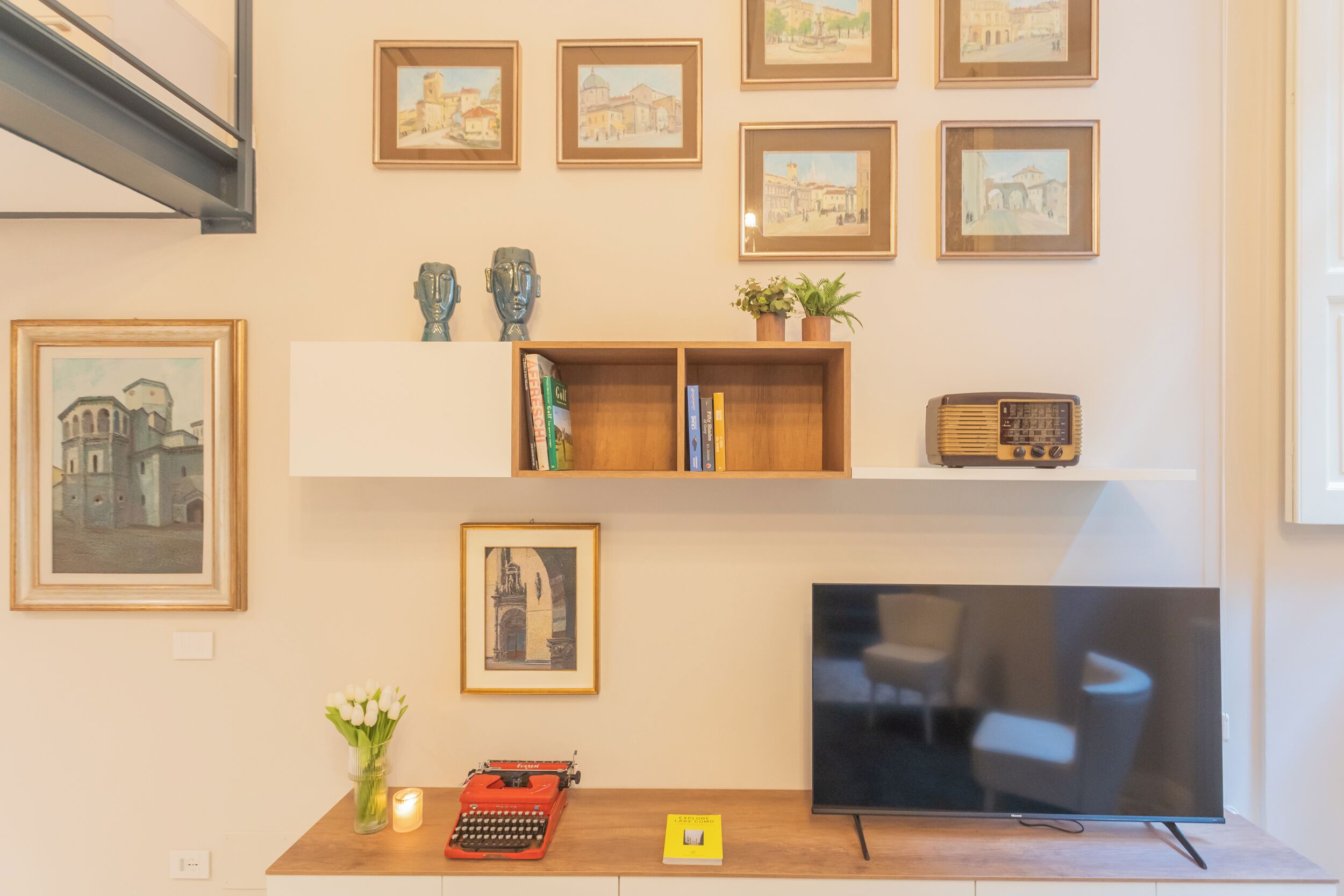 A living room with a flat-screen TV, wooden shelves with books and decorative items, framed pictures on the wall, and a vintage typewriter on a wooden table.