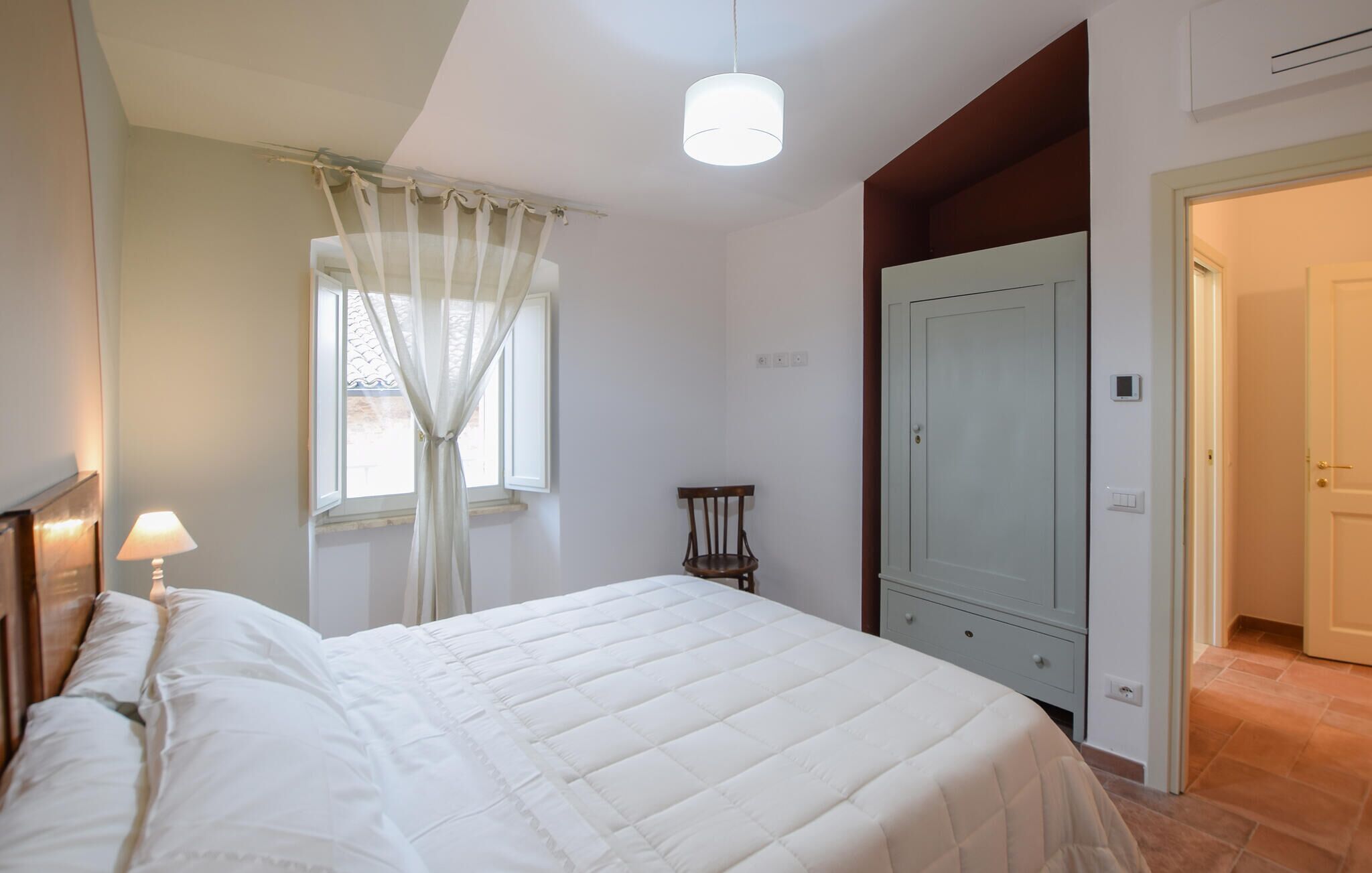 Un dormitorio con cama, una silla, un armario y una ventana con cortinas.