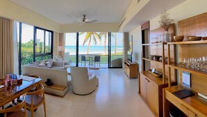 A spacious living area with a balcony overlooking the beach, a dining area, and a wooden shelf with bowls and glasses.