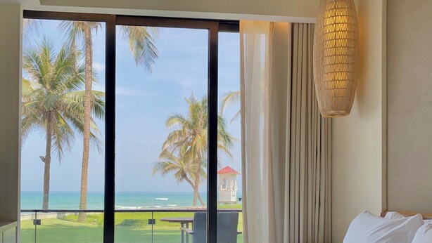 A bedroom with a large bed, a view of the ocean, palm trees, and a beach.