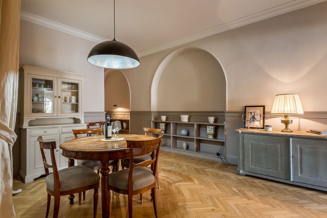 A dining area with a round wooden table, chairs, a hanging light fixture, a cabinet with glass doors, and shelves with decorative items.