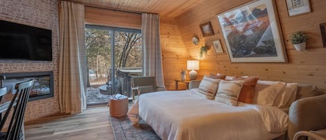 A cozy bedroom with a large bed, a TV mounted on a brick wall, a ceiling fan, and a window with curtains.