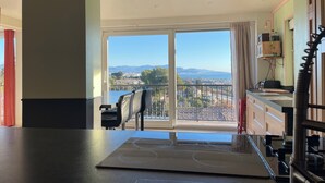 A kitchen with a large island, a dining area with a chair, and a view of a cityscape through a large window. - TOP VILLA PALOMA ANTIBES Vue Mer, Montagne, FIBRE, CLIME, POOL (Antibes)