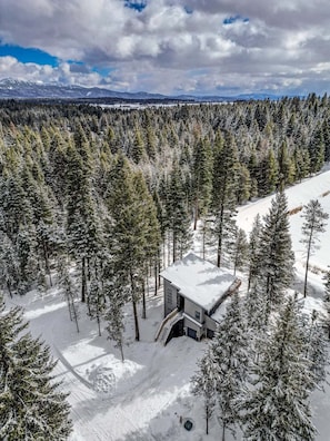 Una cabana plena de neu envoltada d'un bosc dens, amb un cel clar i muntanyes llunyanes.