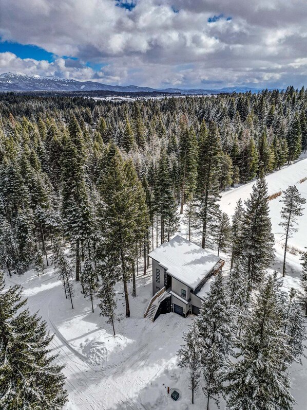 Uma cabana coberta de neve, rodeada por uma floresta densa, com céu claro e montanhas ao longe.