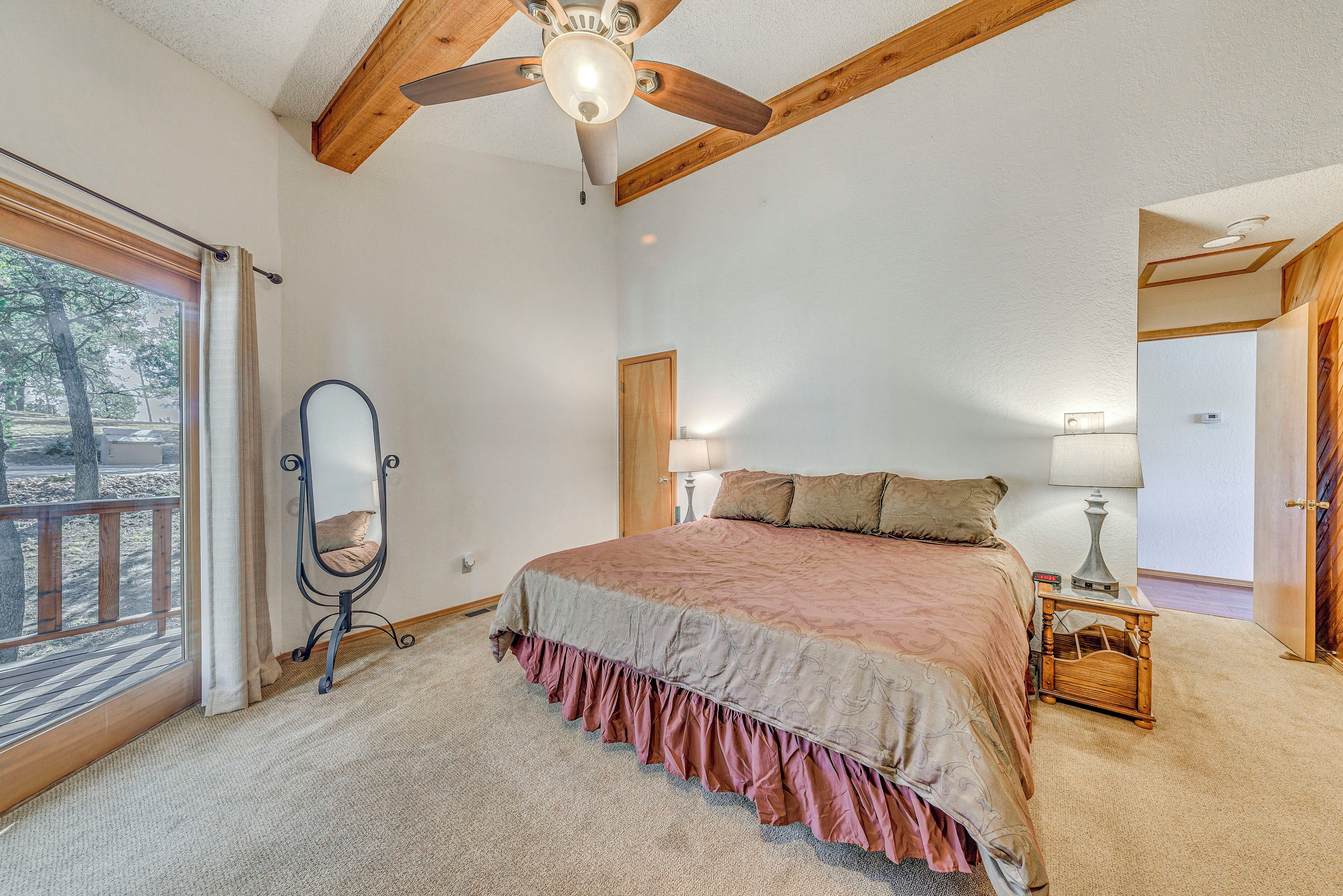Een slaapkamer met een groot bed, houten balken in het plafond, een plafondventilator, een raam met gordijnen, een nachtkastje met een lamp en een spiegel.