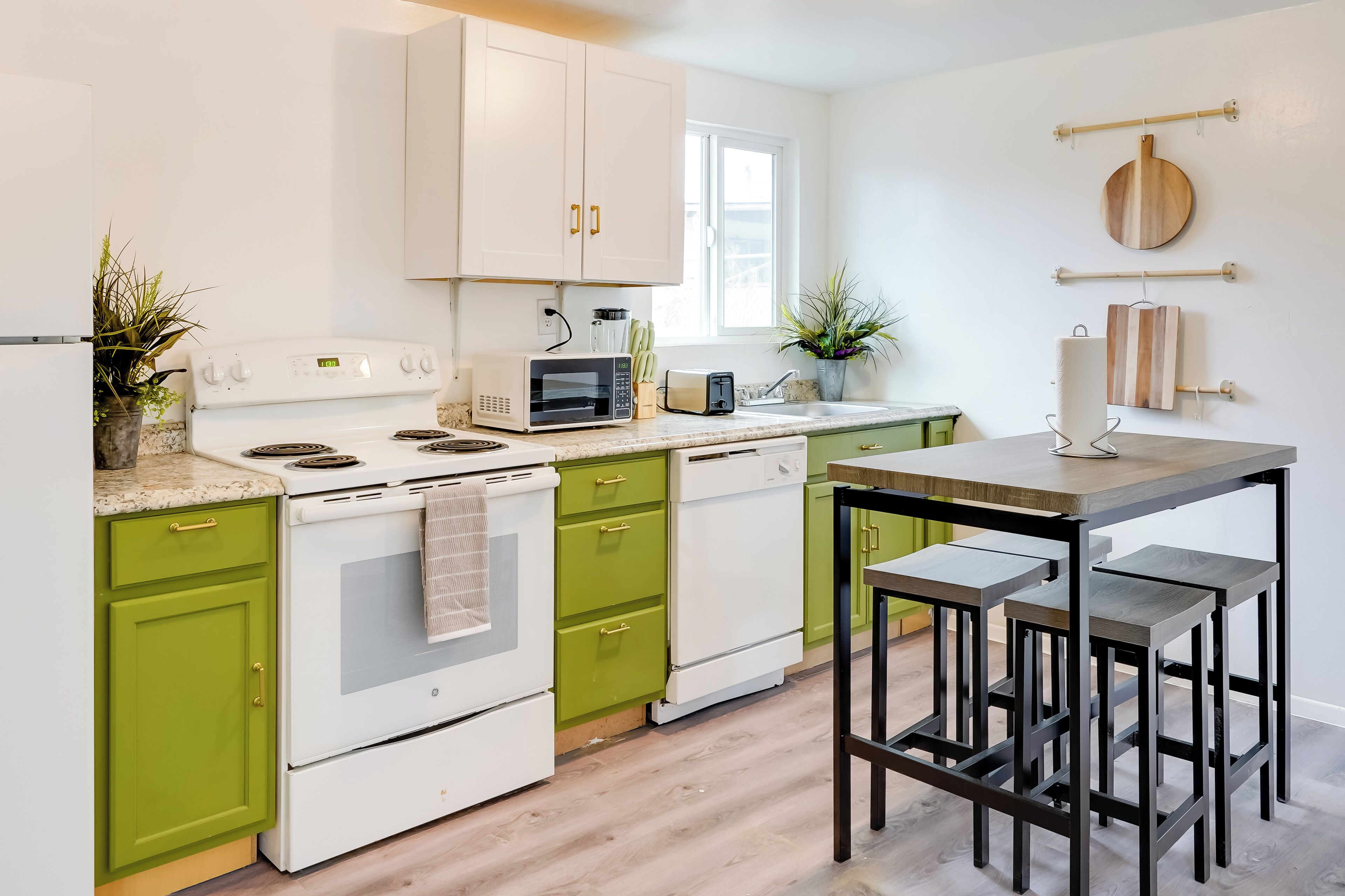 Una cocina moderna con armarios blancos y verdes, microondas, tostadora y una mesa de madera con taburetes.