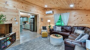 A wooden interior with a sofa, ottoman, and TV. - Explore Lakes & Beaches! Creekside Glen Arbor Home (Glen Arbor)