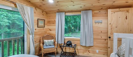 A wooden cabin room with a bed, a wicker chair, a small table with a fan, and a window with curtains.