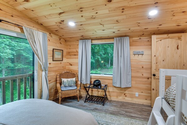 A wooden cabin room with a bed, a wicker chair, a small table with a fan, and a window with curtains. - Explore Lakes & Beaches! Creekside Glen Arbor Home (Glen Arbor)