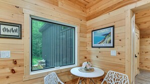 A wooden cabin interior with a large window, two white chairs with patterned cushions, a small table with a vase of flowers, and two framed pictures on the walls. - Explore Lakes & Beaches! Creekside Glen Arbor Home (Glen Arbor)