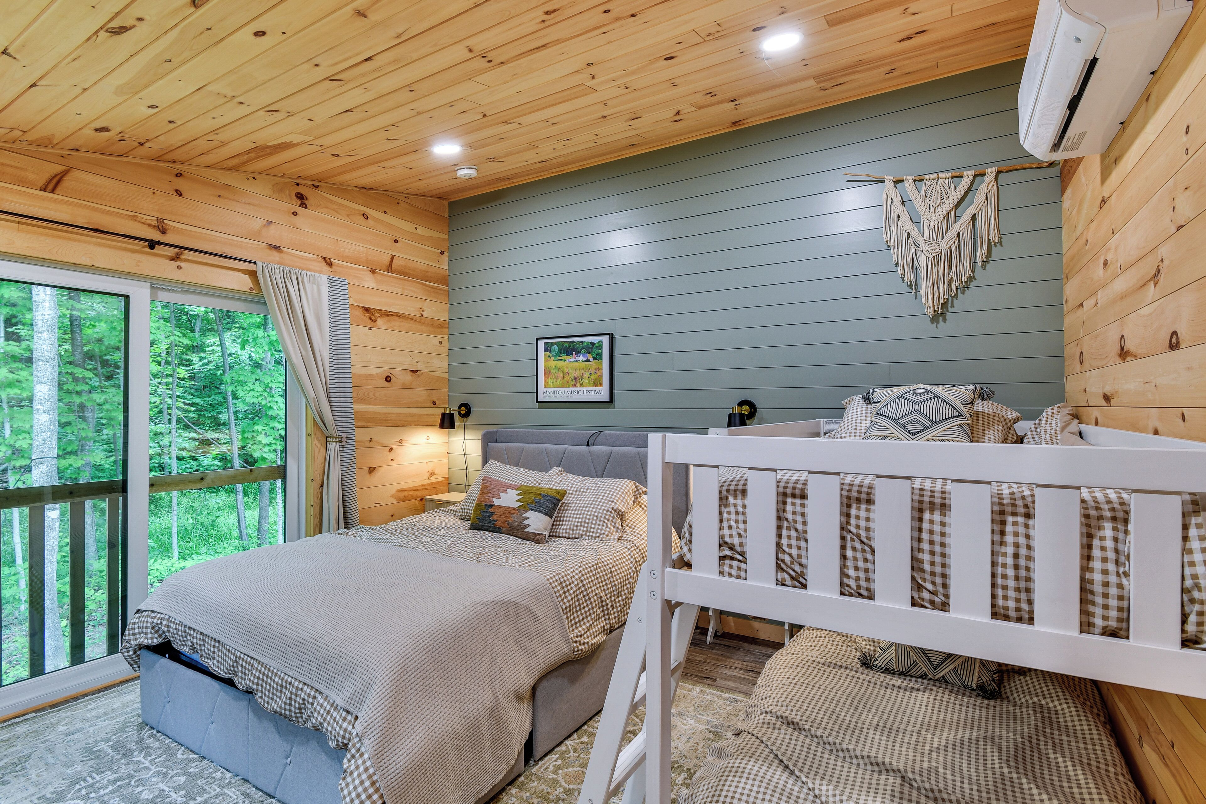 A wooden cabin bedroom with a bed, a crib, a window with curtains, a wall-mounted air conditioner, and a framed picture.