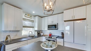 Una cocina moderna con gabinetes blancos, una isla central con un bol de fruta, electrodomésticos de acero inoxidable y una lámpara de araña.