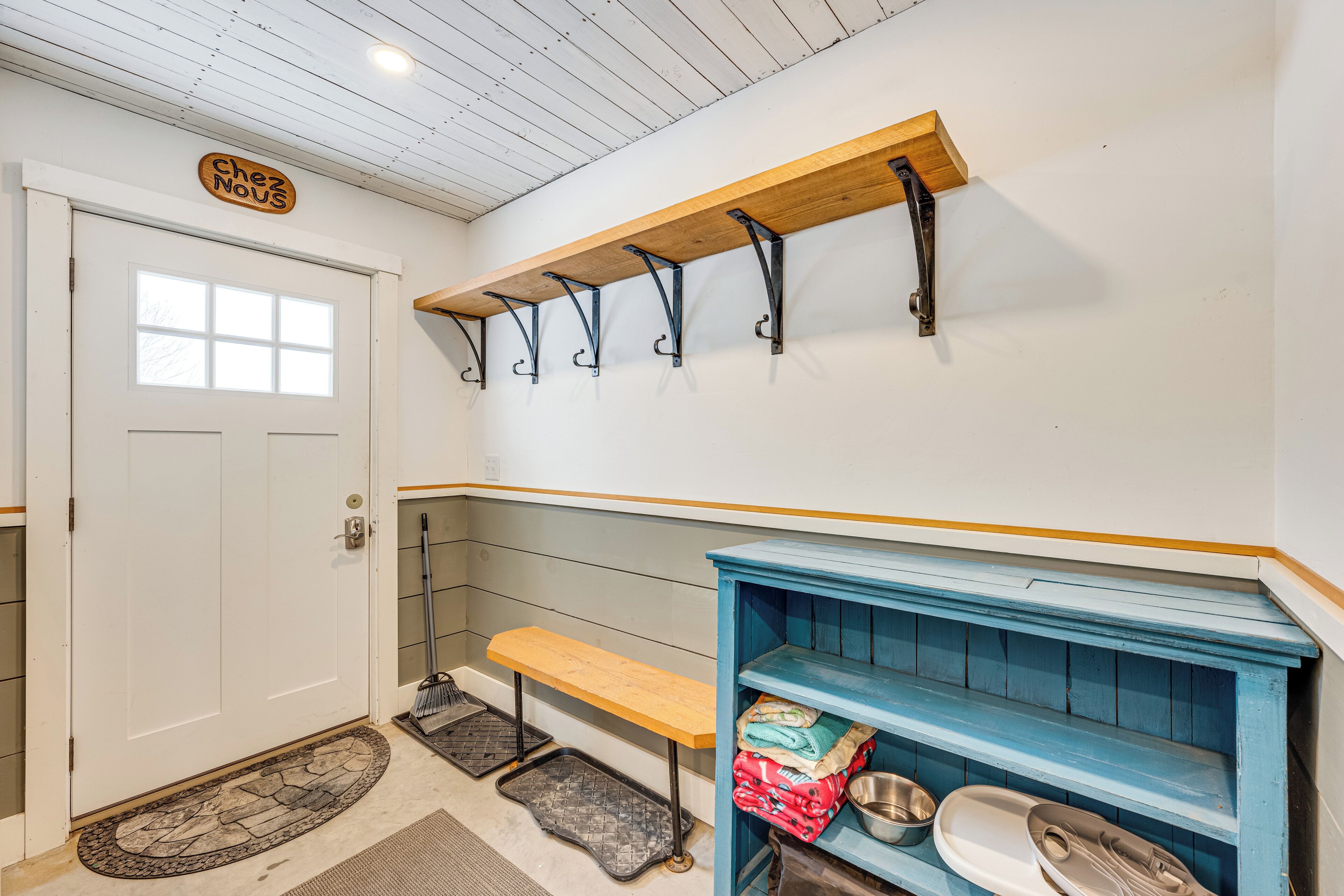 Una habitación con una puerta blanca, un banco de madera, un estante azul con bolsos, y un estante de madera con ganchos.