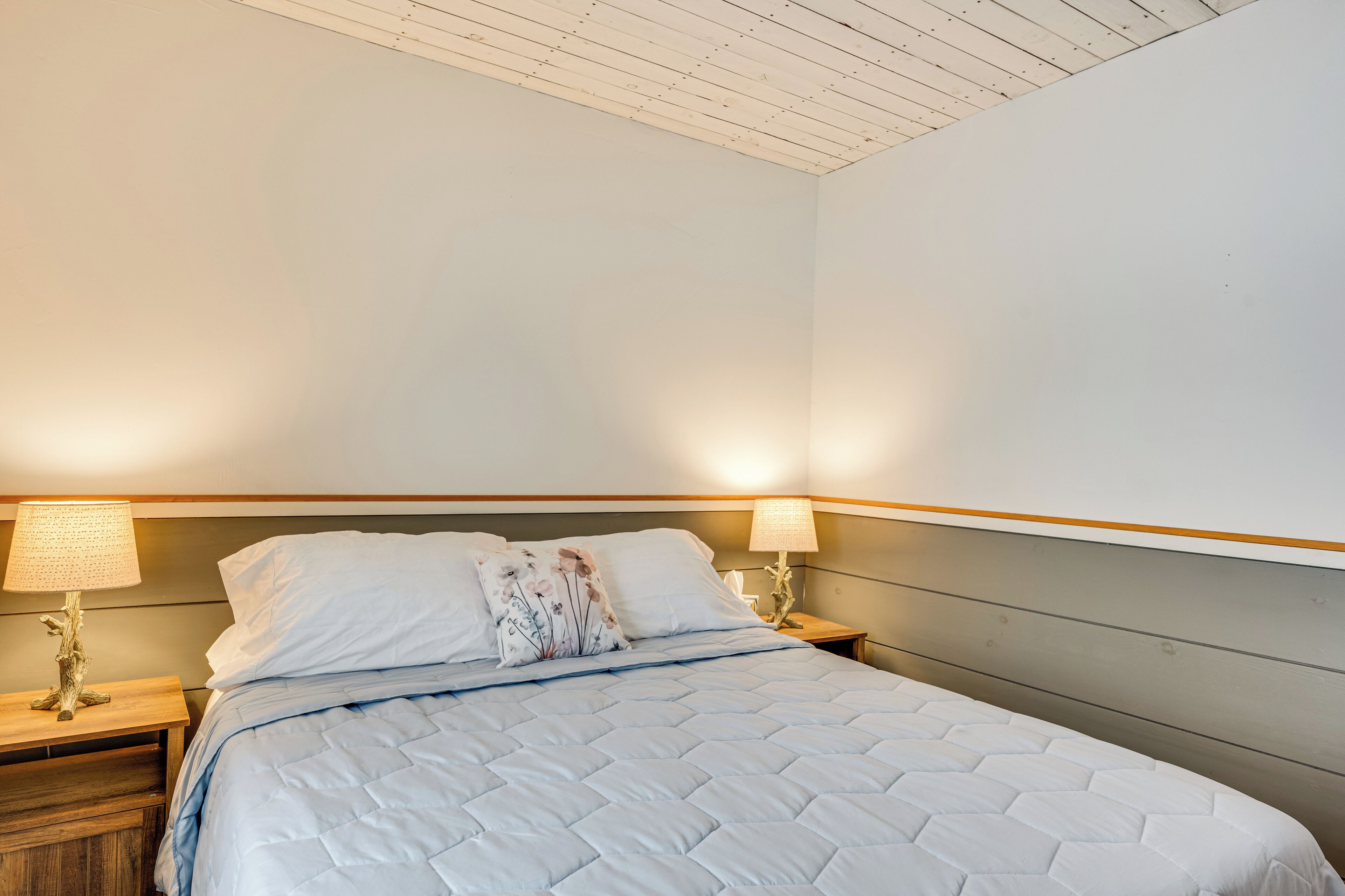 A neatly made bed with a quilted cover, two bedside lamps, and a floral pillow on a wooden nightstand.