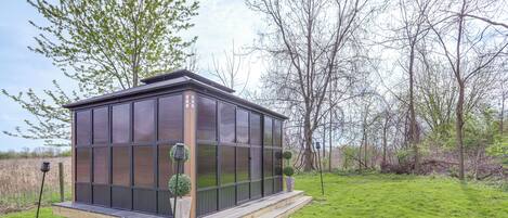 Ein modernes Haus aus Glas mit einer Holzterrasse, umgeben von einer grünen Rasenfläche und Bäumen.