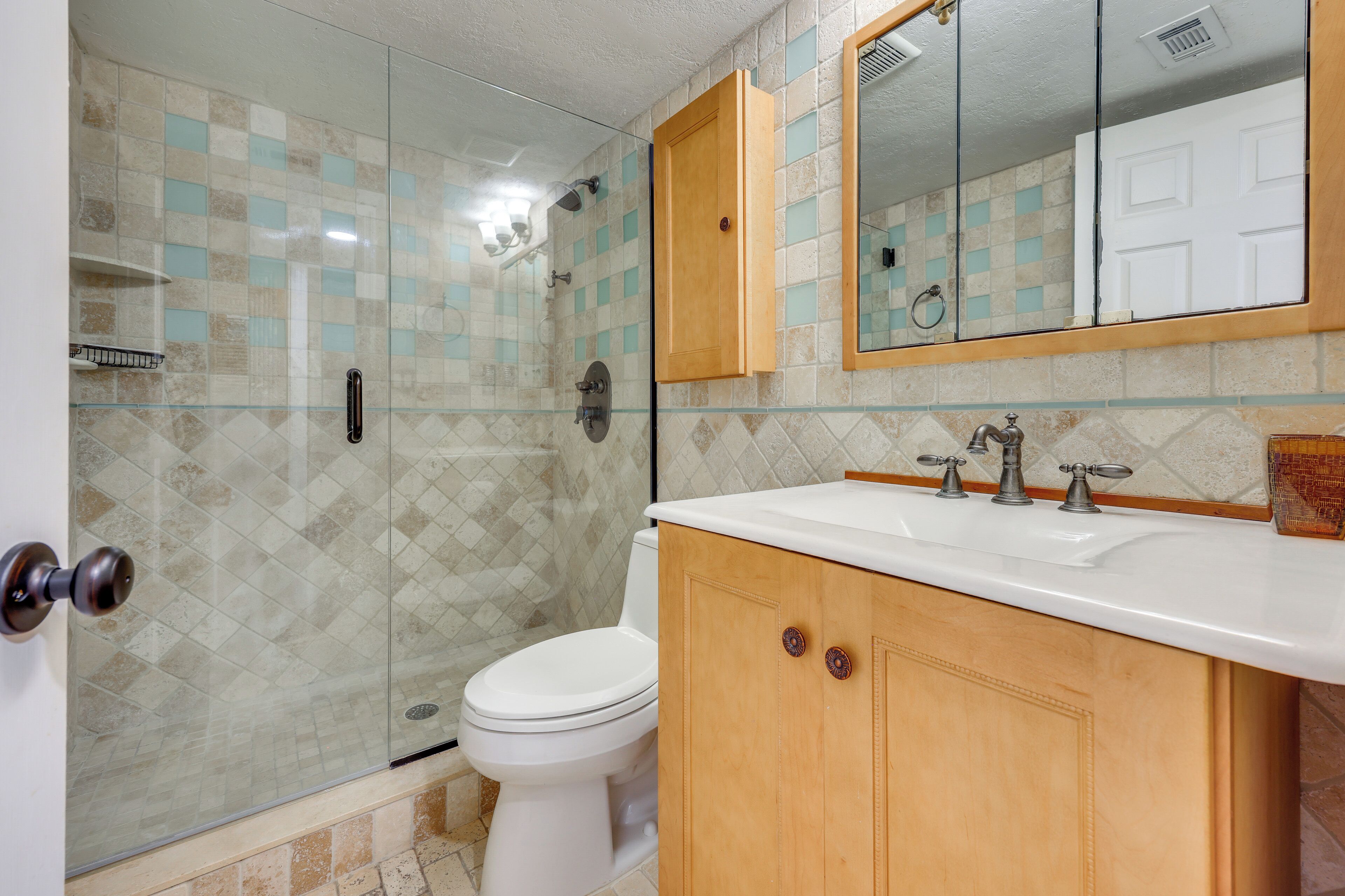Un baño con cabina de ducha de vidrio, un inodoro blanco, un tocador de madera con espejo y un salpicadero de azulejos.