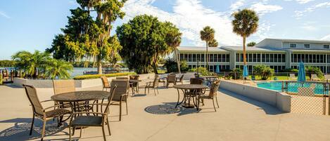 An outdoor patio area with tables and chairs, a pool, and a building in the background.