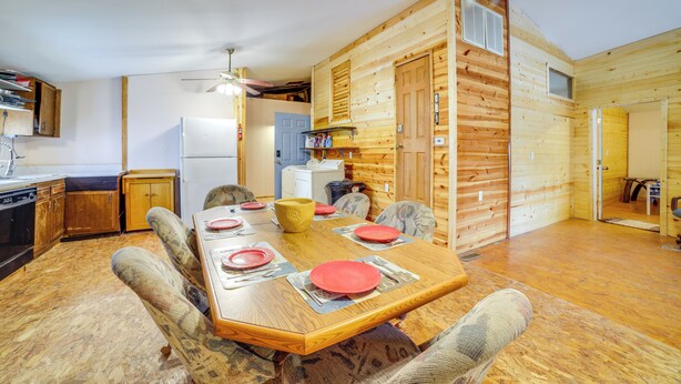 A compact kitchen with a refrigerator, stove, and wooden cabinets. A dining table with chairs and red plates is set up in the center. - Rustic Apartment w/ Sunset Views in Elkhart! (Elkhart)