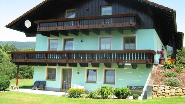 A two-story building with a balcony, a stone wall, and a grassy lawn. - Comfortably furnished vacation apartment with panoramic views (Arnbruck)