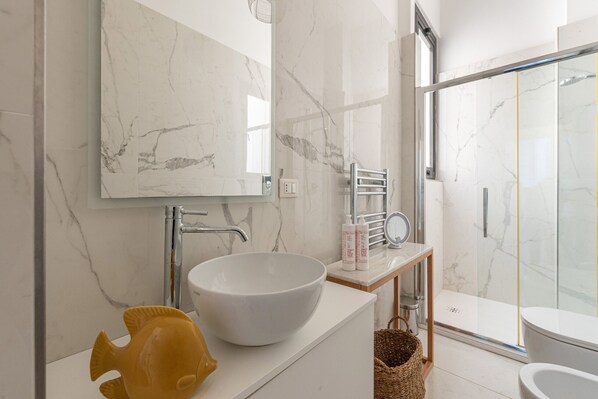 A modern bathroom with a large white basin, a marble wall, a glass shower enclosure, and a wooden vanity.