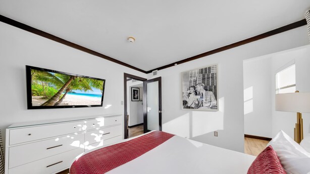 A modern bedroom with a large bed, a television mounted on the wall, and a door leading to another room. - Two Bedroom Suites South Beach (Miami Beach)