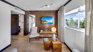 A modern living room with a flat-screen TV mounted on the wall, a sofa, a coffee table, and a view of the outdoors through large windows. - Two Bedroom Suites South Beach (Miami Beach)