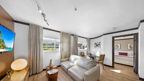 A modern living room with a sofa, a coffee table, and a TV. There is a window with curtains, a dining area, and a bed in the background. - Two Bedroom Suites South Beach (Miami Beach)