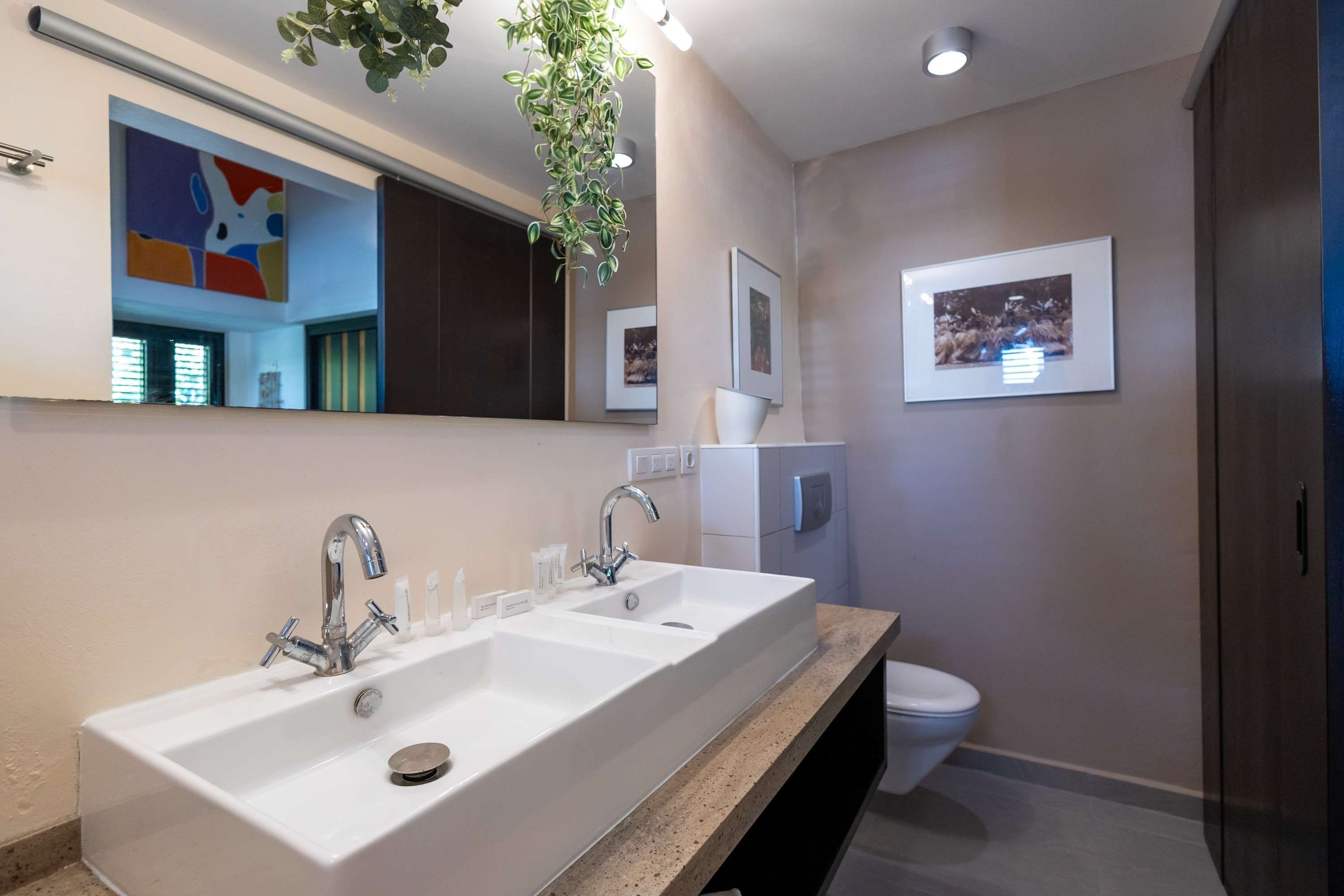 A modern bathroom with a large white sink, two faucets, a mirror, and a toilet.