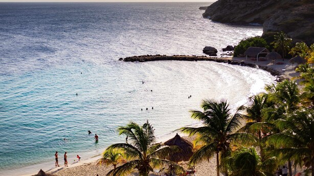 A beach with clear blue water, palm trees, and a rocky cliff. - Indigo Garden at Blue Bay Golf Beach Resort (Sint Michiel)