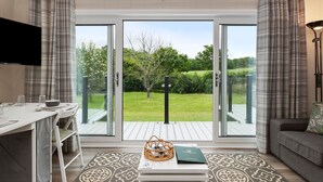 A modern living room with a white sofa, a dining table with chairs, a TV mounted on the wall, and a view of a green garden through large sliding glass doors. - Mallard s Rest - Countryside Hot Tub (Narberth)
