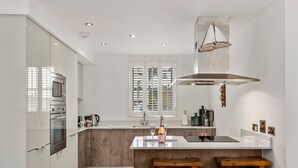 A modern kitchen with a central island, wooden stools, and a large window with shutters. - Maes-y-berllan - Close to Village and Beach (Saundersfoot)