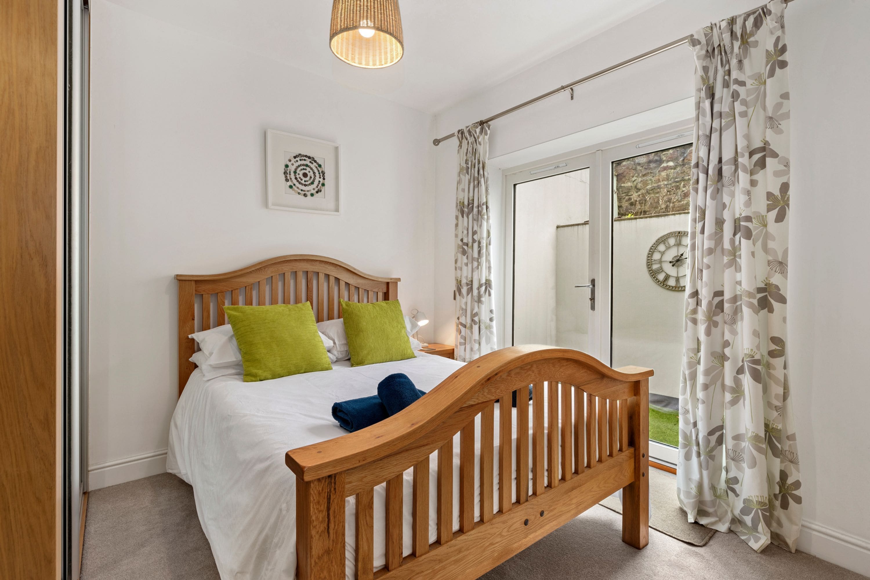 A bedroom with a wooden bed, a bedside table, a wall clock, and a window with curtains.