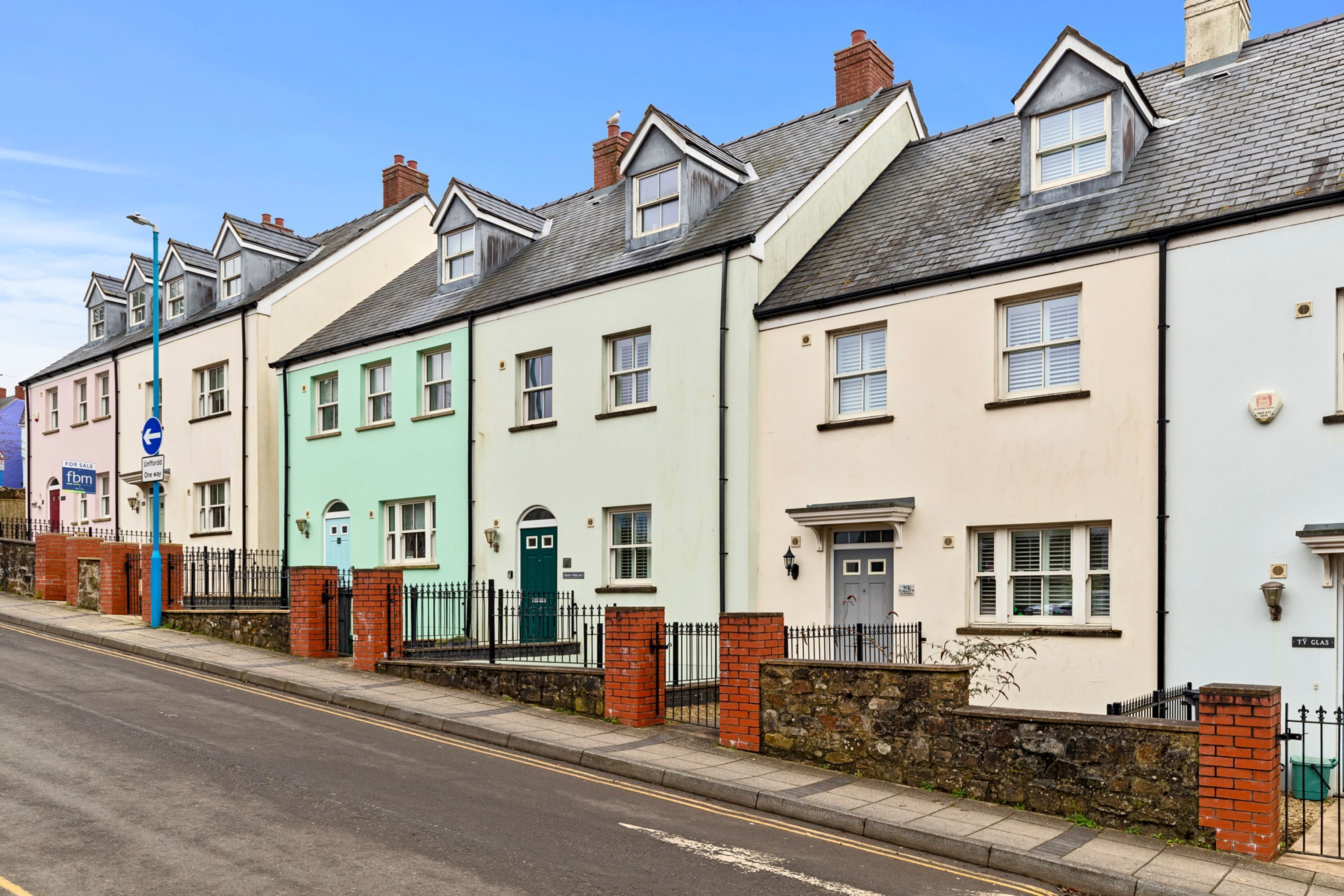 A row of terraced houses with distinct pastel colors, a clear road in front, and a blue sign indicating a nearby location.