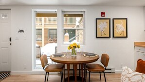 A dining area with a round wooden table, four chairs, and a vase with yellow flowers. There are two framed artworks on the wall, and a fire alarm is mounted above the table.