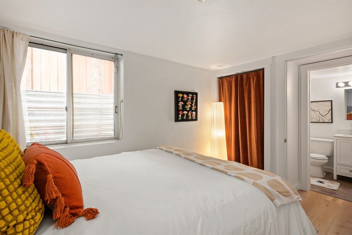 A bedroom with a bed, a window with blinds, a wall-mounted artwork, and a bathroom visible through an open door.
