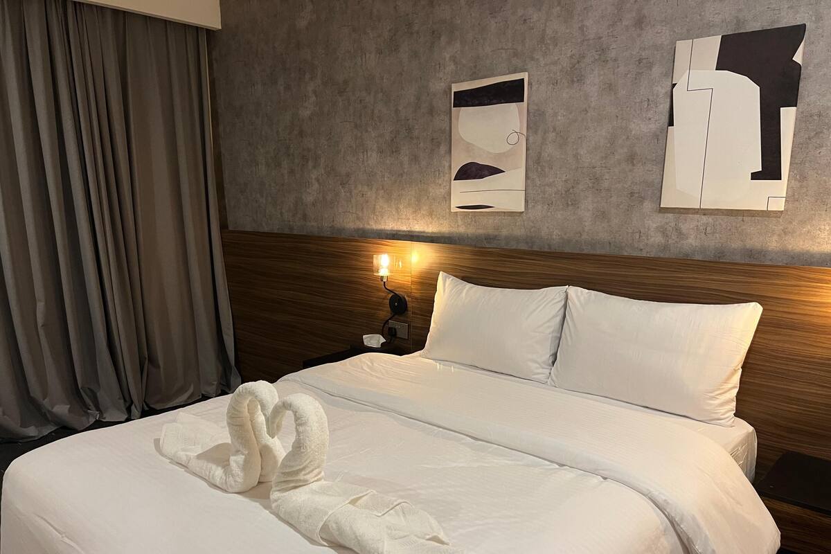 A hotel room with a neatly made bed, a wooden headboard, a nightstand with a lamp, and a wall with abstract art.