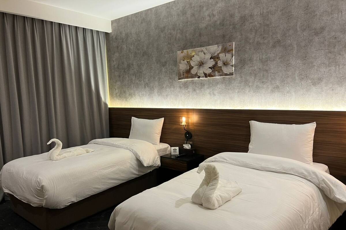 A hotel room with two beds, each with a white swan-shaped towel, a wooden headboard, a wall-mounted light, and a framed floral artwork.