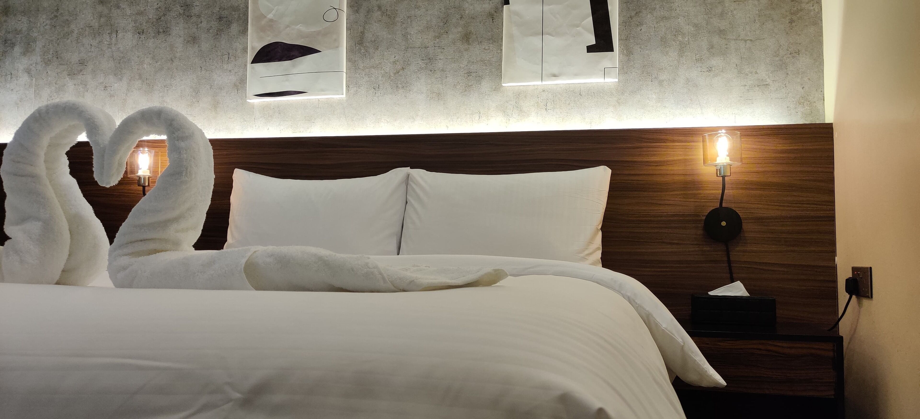 Un lit bien fait, avec du linge de lit blanc, une tête de lit en bois et une table de chevet surmontée d’une lampe.