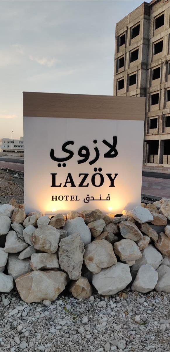 Un letrero de LAZÔY HOTEL con texto en árabe, sobre una superficie de grava con rocas al frente, y un edificio en el fondo.