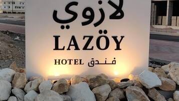 Un letrero de LAZÔY HOTEL con texto en árabe, sobre una superficie de grava con rocas al frente, y un edificio en el fondo.
