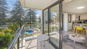 A balcony with a glass door, a dining area with yellow chairs, and a view of trees and a swimming pool. - Tasman Towers 4 - Conveniently located in the CBD (Port Macquarie)