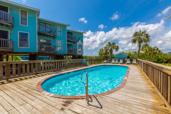 A swimming pool with a wooden deck, surrounded by a wooden fence, and adjacent to a turquoise building with balconies. - The Sands 107 (Gulf Shores)