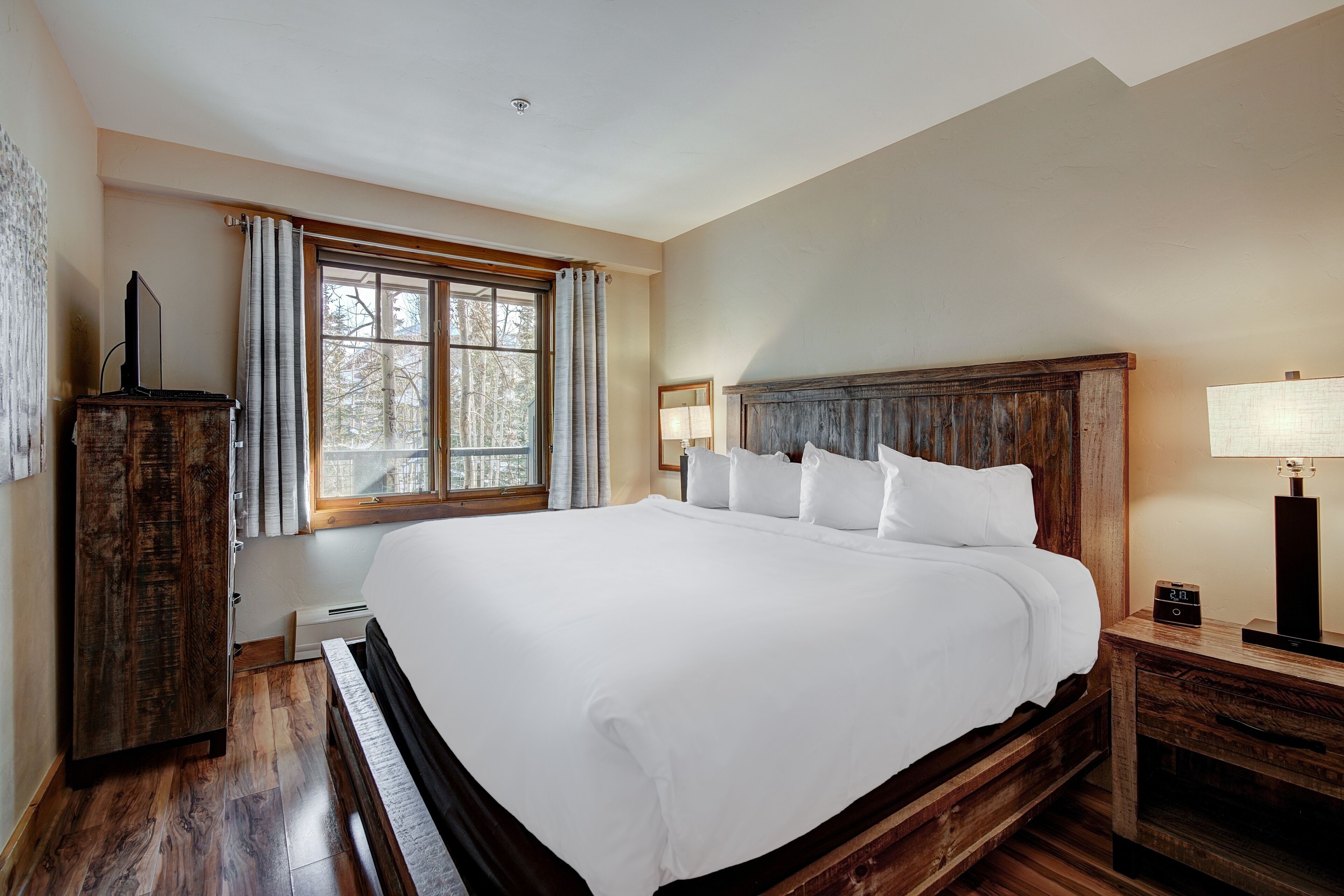 A large bed with white linens, a wooden headboard, a nightstand with a lamp, and a window with curtains.