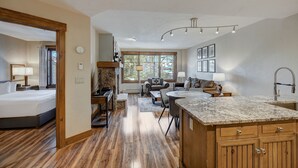 A spacious hotel room with a large bed, a sitting area with a sofa and coffee table, a kitchen with granite countertops, and a view of the outdoors through large windows. - Cozy Ski Chalet With Large Balcony 1 Bedroom Condo (Breckenridge)