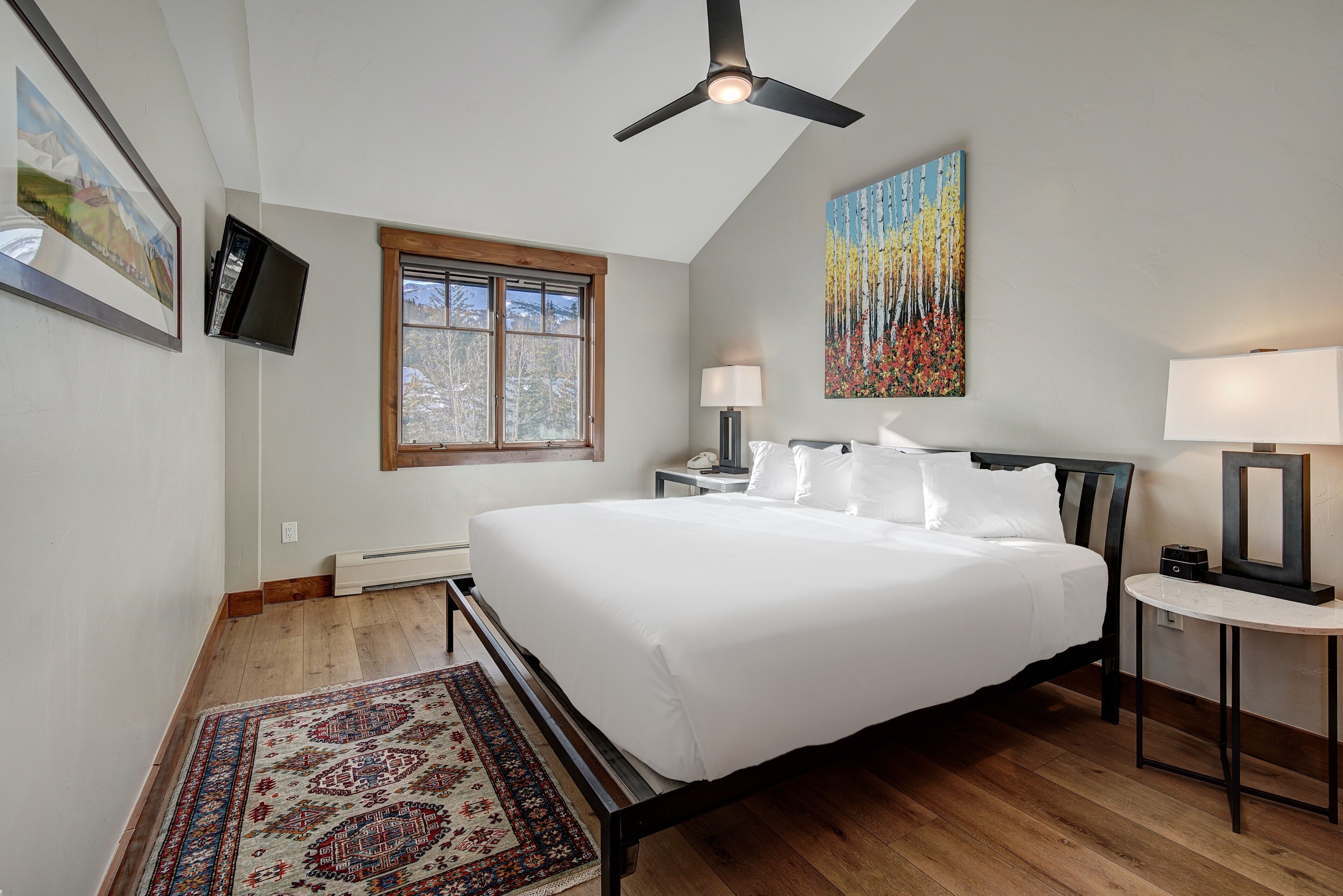 A bedroom with a large bed, a ceiling fan, a window, a TV, and artwork on the walls.